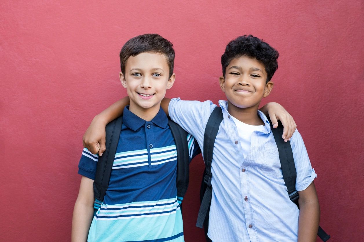 Best children friends standing with hand on shoulder against red background. Happy smiling classmates standing together on red wall after school. Portrait of multiethnic schoolboys enjoying friendship. (Best children friends standing with hand on shou